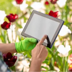 Image of gloved hand on one side and a hand using a tablet device with flowers in the background